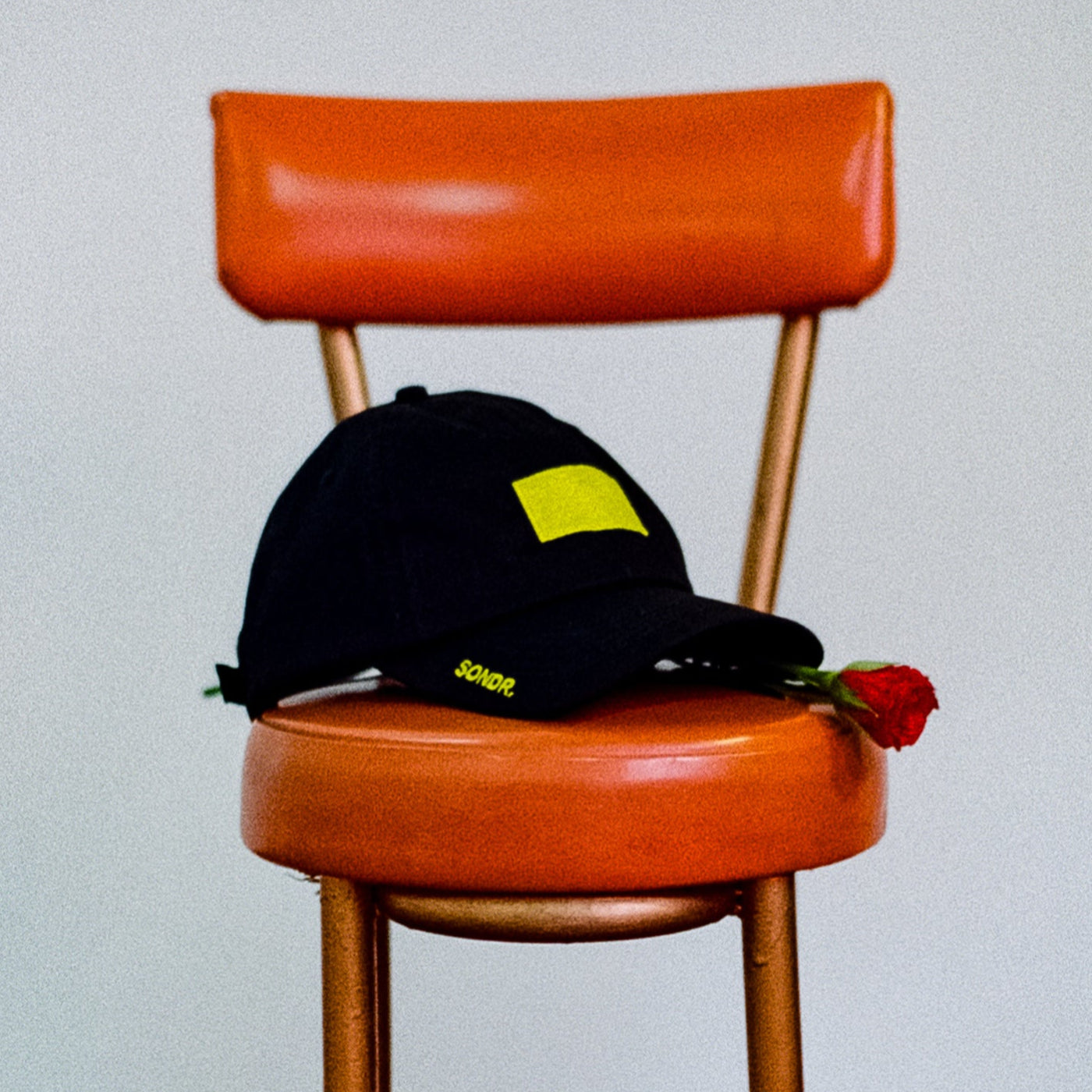 Vintage burnt orange bar stool in a room alone with black SONDR. Obscure Sorrows Dad Hat & single rose on the seat