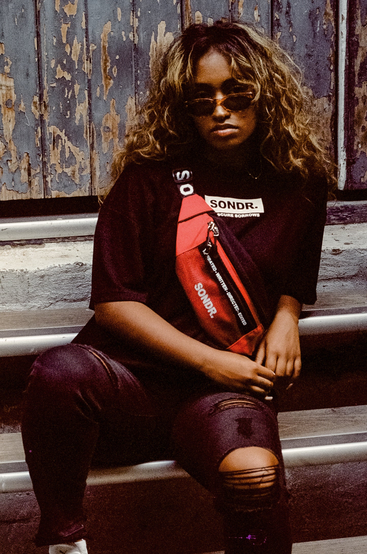 Model wearing Red Cross Body Bag sitting on stairs wearing SONDR. T-Shirt