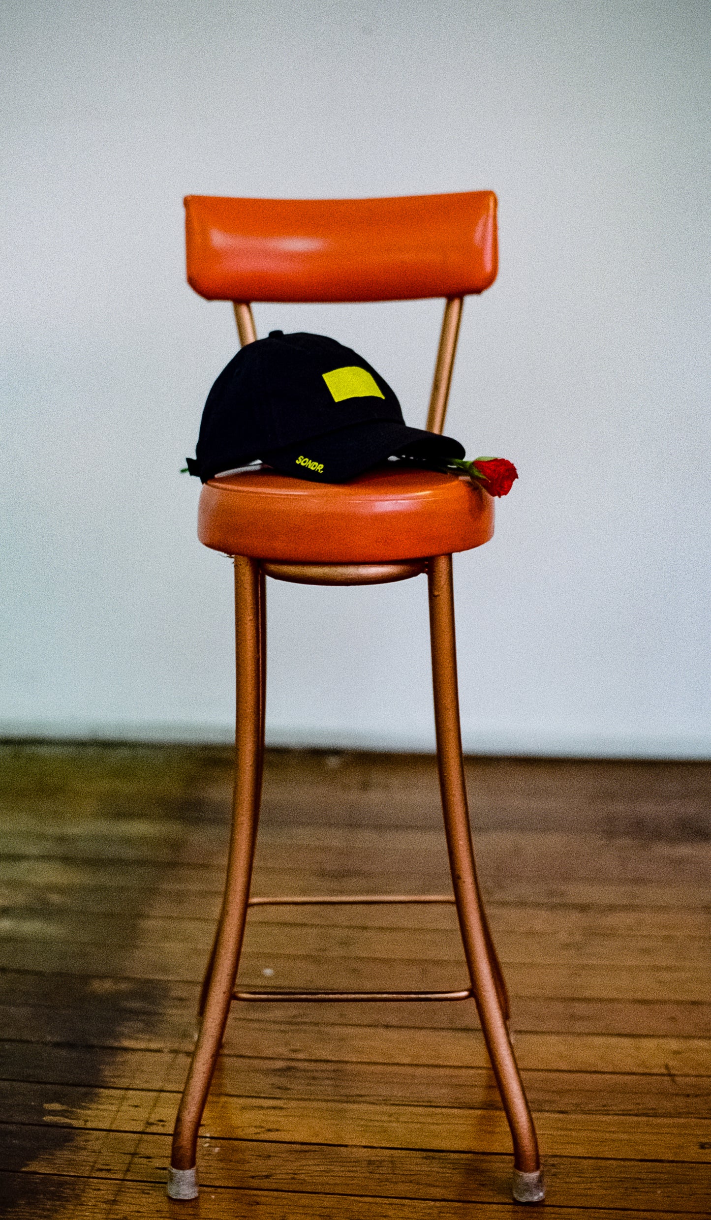 Vintage burnt orange bar stool in a room alone with black SONDR. Obscure Sorrows Dad Hat & single rose on the seat